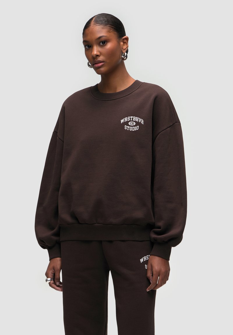 Felpa marrone con colletto a girocollo e vestibilità rilassata, caratterizzata da un logo ricamato bianco "WESTBHVR STUDIO" sul petto. Tessuto morbido.
