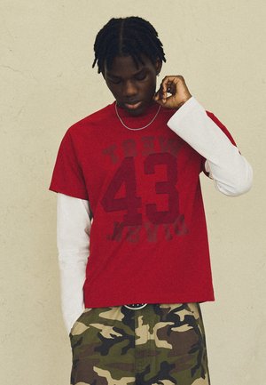 Jonge man met gevlochten haar die een rood "43" shirt draagt over witte lange mouwen en camouflagebroek, staand tegen een effen muur.