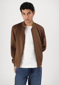 Brun bomberjakke med et struktureret mønster, ribbet krave og manchetter, frontlynlås, båret over en hvid t-shirt, parret med blå jeans.