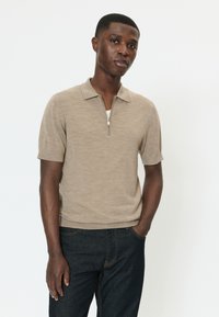 Junger Mann, der einen beigefarbenen Kurzarmpullover mit Reißverschluss am Kragen und eine dunkle Jeans trägt, mit einer Hand in der Tasche stehend vor weißem Hintergrund.