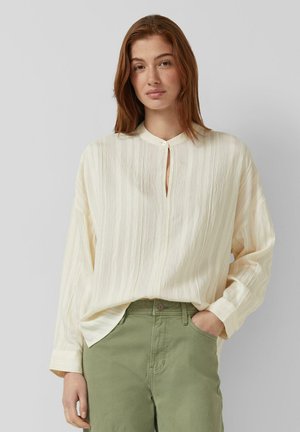 Femme aux cheveux roux portant un chemisier crème texturé sans col et un pantalon vert, debout avec une main dans la poche sur un fond gris.