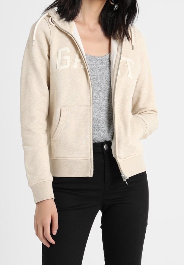 Donna che indossa una felpa beige con zip e lettering "GANT", una maglietta grigia sotto e pantaloni neri, in piedi contro uno sfondo bianco.