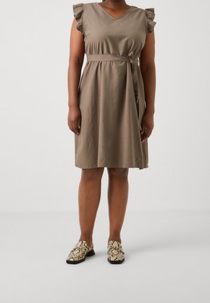 Vrouw draagt een taupekleurige jurk tot op de knie met gerimpelde mouwen en ceintuur, gecombineerd met beige en zwarte loafers met slangenprint, staand op een grijze vloer.