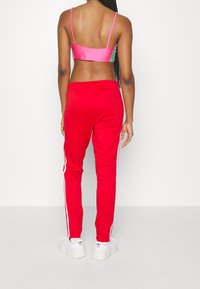 Rote Sporthosen mit weißen Seitenstreifen, kombiniert mit einem pinken satin Crop-Top. Das Model hat geflochtenes Haar und sieht von der Kamera weg.