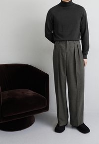 Czarny golf i szare plisowane spodnie o gładkiej fakturze. Czarne kapcie. W tle brązowy aksamitny fotel o zaokrąglonym kształcie.