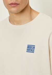 Beige bavlněné tričko s kulatým výstřihem a malým modrým logem "AMERICAN VINTAGE" na levé části hrudi. Hladká textura.