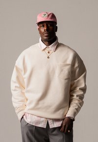 Kremfarget oversized sweatshirt med brystlomme, lagt over en rosa stripete skjorte. Rosa caps med et sirkulært logo, gullsmykke detaljer.
