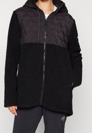 Person trägt eine schwarze Jacke mit Reißverschluss, gesteppter Oberteil und Kapuze, kombiniert mit dunklen Hosen mit Mammut-Logo.