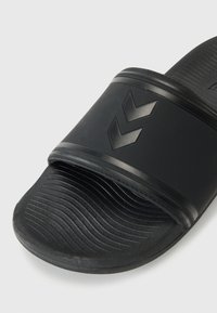 Black slide sandal with a textured surface, featuring a wide strap and raised chevron logo. Flat, ridged sole for grip and comfort.