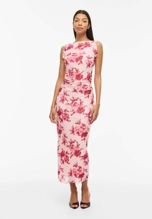 Mujer con vestido midi sin mangas de color rosa con estampado floral rojo y detalles de volantes en los hombros, de pie con zapatos negros de tacón abierto sobre fondo blanco.