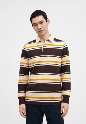 Jeune homme portant un polo à manches longues rayé marron, crème et jaune, debout devant un fond blanc uni.