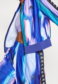 Persona che indossa una giacca e pantaloni della tuta Kappa dai colori vivaci blu e viola, con un reggiseno sportivo lilla, addome scoperto, in una posa dinamica.