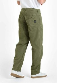 Persoon in olijfgroene ontspannen broek met opgestikte zakken, een zwart logo op de rechter achterzak, witte sneakers en een wit shirt.