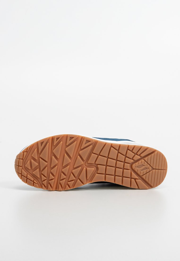 Semelle d'une chaussure de sport en caoutchouc marron avec des motifs de bande de roulement triangulaires et rectangulaires sur un fond blanc.