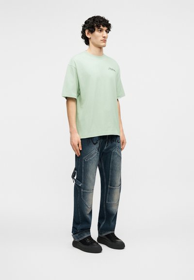 Jeune homme debout portant un t-shirt vert clair oversize et un pantalon cargo en jean foncé délavé avec des baskets noires sur un fond uni.