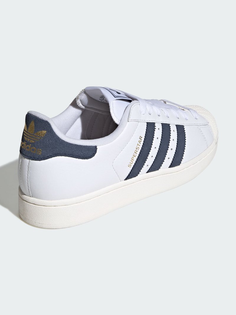 White Adidas Superstar sneaker with black stripes, gold "SUPERSTAR" text on side, and black heel tab with gold Adidas logo.