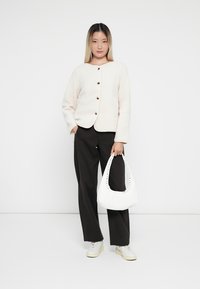 Veste blanche texturée avec un col rond et une fermeture à boutons. Pantalons noirs à jambes larges. Sac à main blanc avec des accents cloutés. Baskets blanches.