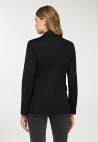 Blazer nero su misura con design strutturato, colletto a revers e maniche lunghe, realizzato in tessuto liscio. Vista posteriore che mostra una silhouette slanciata.