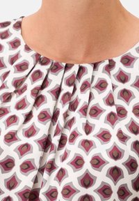 Décolleté et partie supérieure du thorax d'une femme portant une blouse blanche avec un motif géométrique rose et marron et des plis de tissu froncés au col.