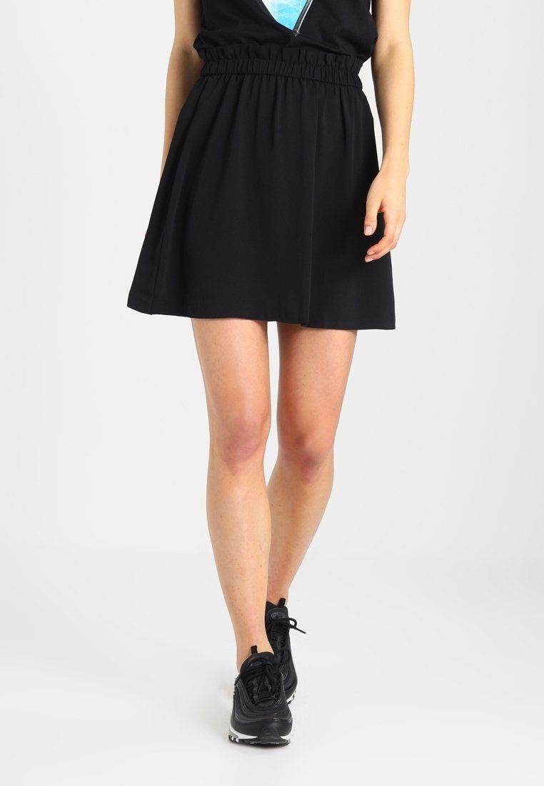 Jupe patineuse noire avec une taille élastique, tissu lisse, design jusqu'aux genoux, associée à des chaussures de sport noires.