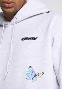 Hellgrauer Hoodie aus weichem Stoff, mit einem schwarzen gestickten Logo auf der Brust und einer bunten Schmetterlingsgrafik an der Seite.