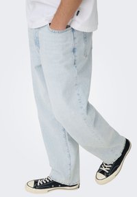 Φωτεινό γαλάζιο τζιν με χαλαρή εφαρμογή, με πολλές τσέπες και διακριτική ξεθώριαση. Συνδυασμένο με μαύρα αθλητικά παπούτσια με λευκές λεπτομέρειες.