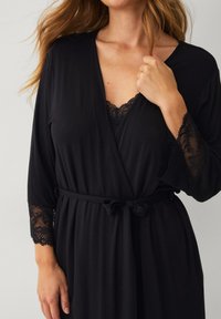 Robe noire enroulée faite d'un tissu doux avec des accents en dentelle sur les manches trois-quarts et une taille nouée. Présente un délicat décolleté en dentelle.