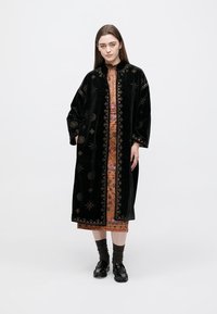 Μαύρο βελούδινο παλτό με περίτεχνη κεντητική, ανοιχτό μπροστά και φαρδιά μανίκια. Φορεμένο πάνω από φόρεμα με σχέδια, συνοδευόμενο από σκοτεινές κάλτσες και παπούτσια.