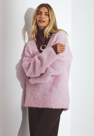 Lichtroze oversized trui gemaakt van fluffy materiaal, met verlaagde schouders en geribde manchetten, gecombineerd met een donkerbruin topje eronder.