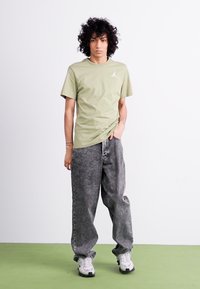 Olivengrøn bomulds t-shirt med et lille hvidt logo, parret med løstsiddende grå denim jeans og hvide sneakers på en grøn gulv.
