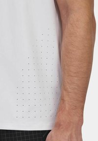 Maglietta sportiva bianca con superficie texturizzata e piccole perforazioni sul lato sinistro, con pantaloni corti neri a contrasto parzialmente visibili.