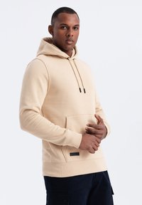 Felpa beige realizzata in tessuto morbido, con tasca frontale, corde con punte nere e un piccolo etichetta nera all'orlo.