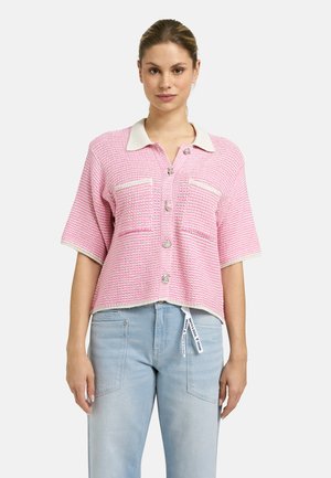 Woman wearing a pink short-sleeve knit cardigan with cream collar and buttons, paired with light blue denim jeans.