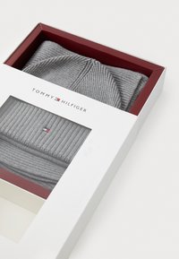 Beanie gri cu dungi, aflat într-o cutie albă cu interior roșu, având un mic logo roșu, alb și albastru în față.