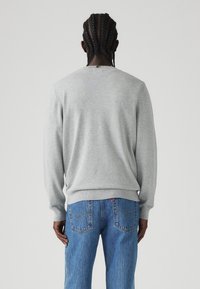 Pilkas megztinis su apvalia iškirpte, su raukšlėtomis apykaklėmis ir hemu, vilkimas virš mėlynų džinsų su išskirtiniu užpakalinių kišenių siuvinėjimu ir etikete.