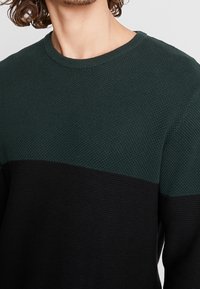 Pull en tricot présentant une section supérieure texturée de couleur vert foncé et une section inférieure noire unie, avec un col rond classique.