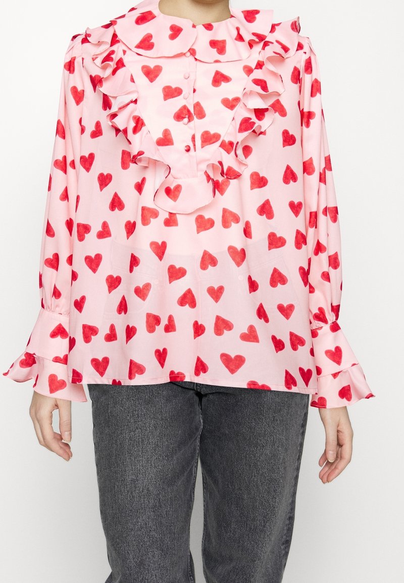 Blouse rose à manches longues avec motif de cœurs rouges, col à volants et plastron devant, portée avec un jean gris foncé.