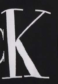 Schwarzer Strickpullover mit einem großen weißen "K" und kontrastierenden strukturierten Akzenten. Der Stoff ist fest gewebt mit sichtbaren Nähdetails.