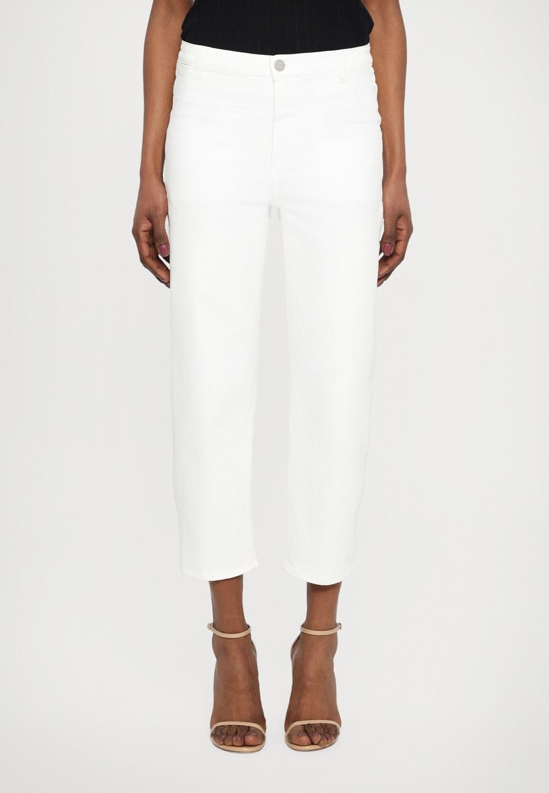 Femme portant un pantalon blanc court taille haute avec fermeture à boutons, associé à des talons nude à bride cheville et un top noir.