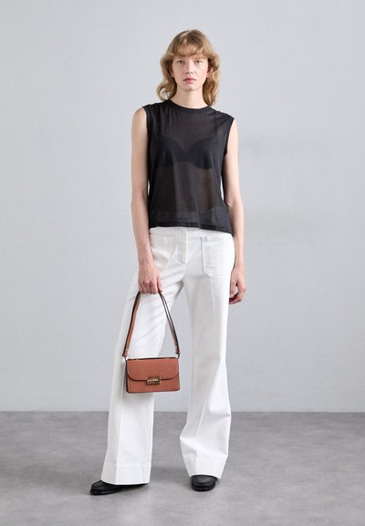 Modelul stă în fața unui fundal gri, purtând un top see-through negru fără mâneci și pantaloni albi largați, ținând o mică poșetă maro.