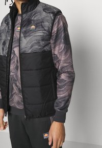 Pufferväst med en marmorerad grå och svart design, med en svart nederdel och dragkedjestängning. Logotyper är placerade på bröstet.