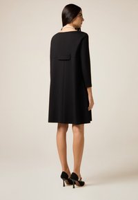 Czarna sukienka typu shift z długimi rękawami, zaokrąglonym dekoltem z tyłu i małą kokardką z tyłu. Uzupełniona czarnymi szpilkami.