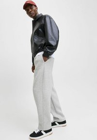 Jacket de piele negru tip bomber, pantaloni de trening gri și adidași negri cu accente albe. Modelul poartă o șapcă roșie, având mâinile în buzunare.