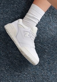 Weiße Ledersneaker mit durchlochter Textur, dicke Gummisohle und minimalistischem Design. Getragen mit einem gerippten weißen Socken auf einem blauen Teppich.