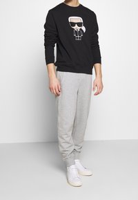 Felpa nera con grafica di un personaggio con occhiali da sole, abbinata a pantaloni grigi chiari e scarpe da ginnastica bianche. Design semplice e informale.