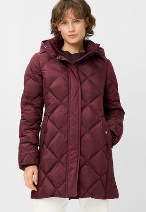 Woman wearing a long, quilted, dark purple winter coat with a hood, standing against a plain light background.