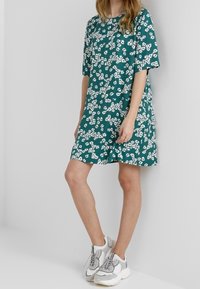 Vestido corto de manga corta en color azul verdoso con un patrón floral blanco, de corte holgado y cuello redondo; combinado con zapatillas en gris y blanco.