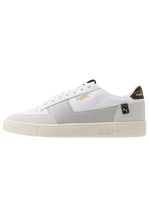 Baskets basses Puma blanches et grises avec logo doré, languette noire au talon et semelle crème texturée avec marquage Puma Shock Wedge.