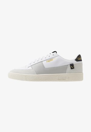 Baskets basses Puma blanches et grises avec logo doré, languette noire au talon et semelle crème texturée avec marquage Puma Shock Wedge.
