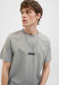 Gray cotton t-shirt with a crew neck and short sleeves. Features a black logo printed on the chest and worn with a silver chain necklace.